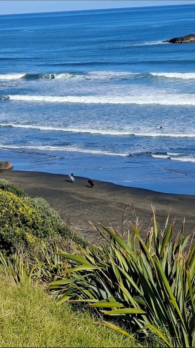 Surfers walking the black sand as lines of swell roll in